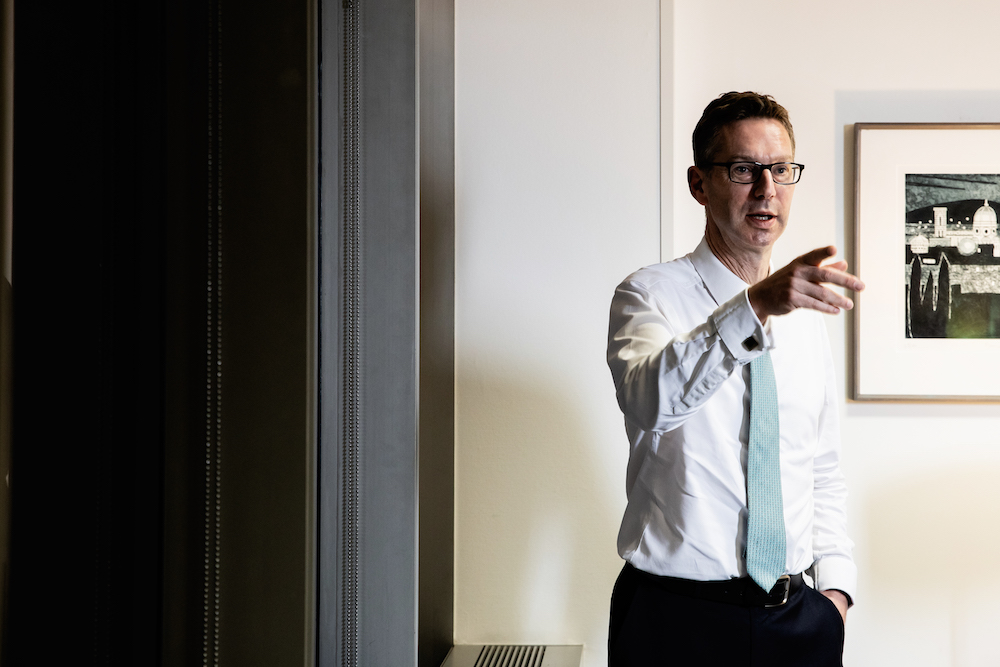 A candid shot of Peter Schofield in an office, pointing at something out of shot, a painting on the wall behind him